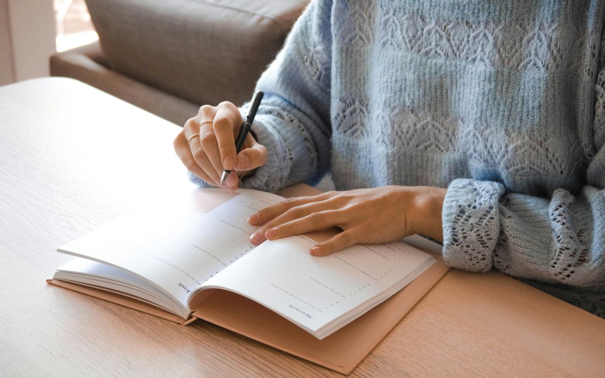 Person writing in an open notebook while sitting at a table.