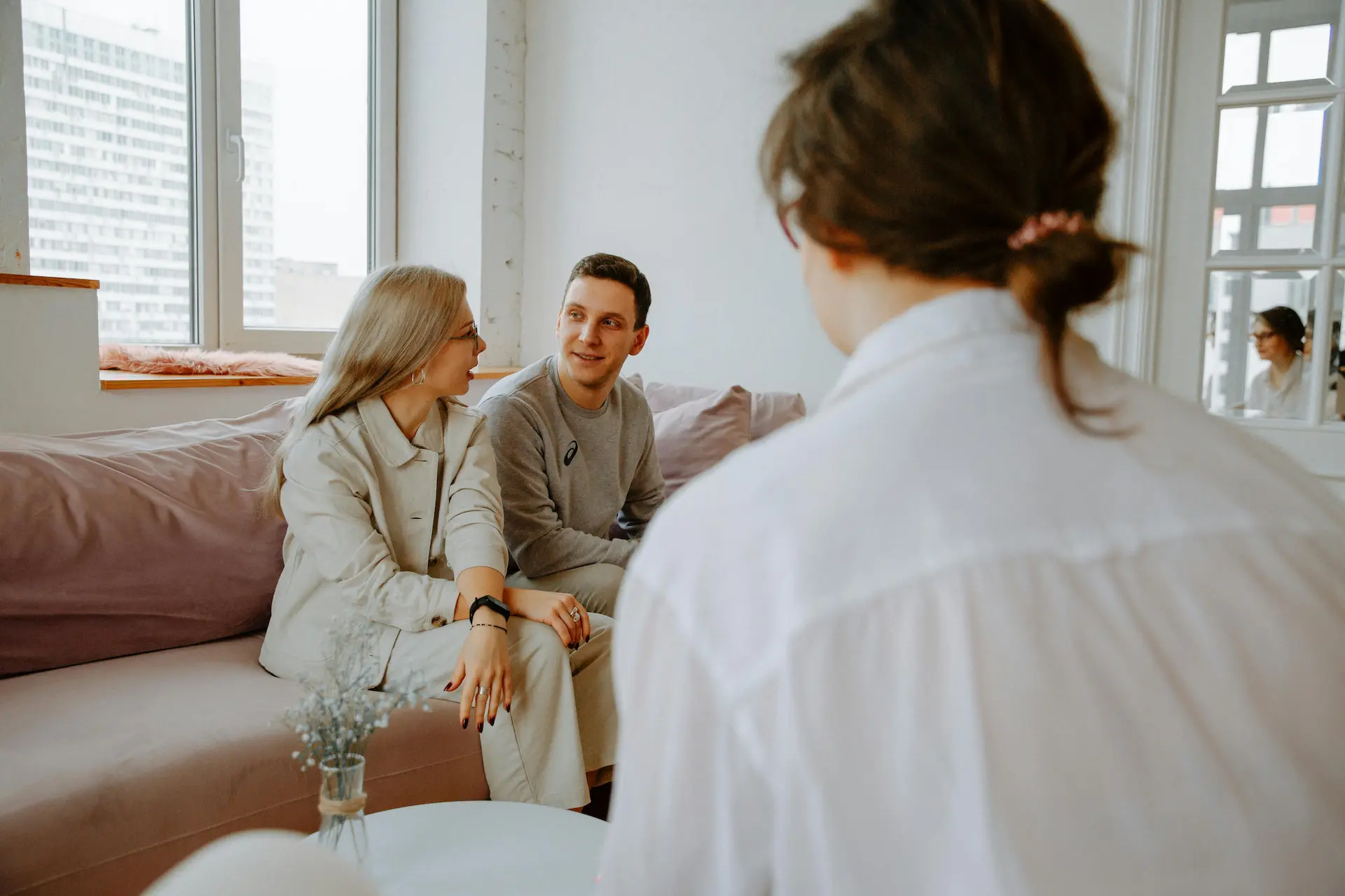 A young couple sits on a sofa talking with a clinical professional in a bright, modern office during a support session.