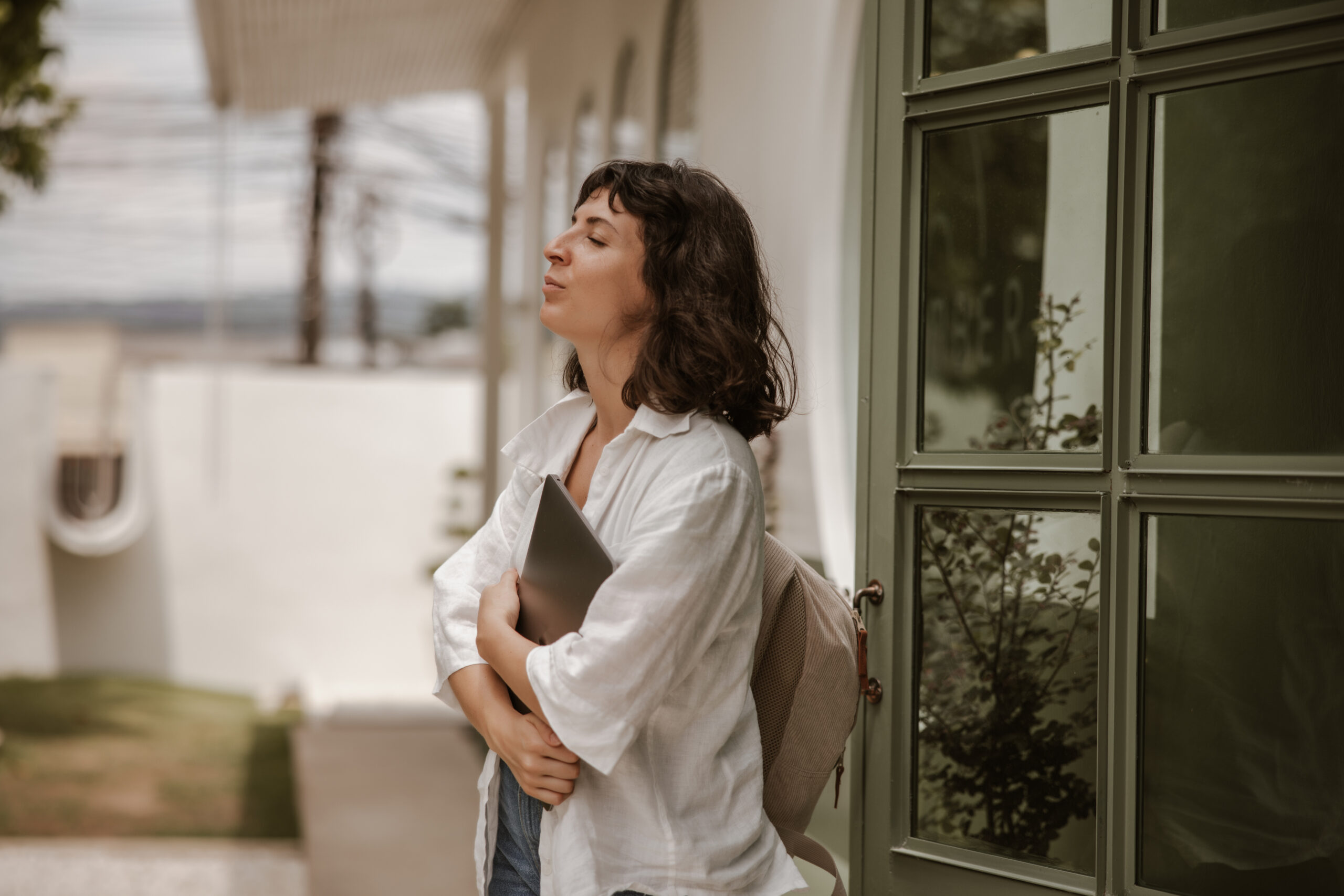 A young woman with a backpack holds a laptop while standing outside a building with her eyes closed in a mindful moment.