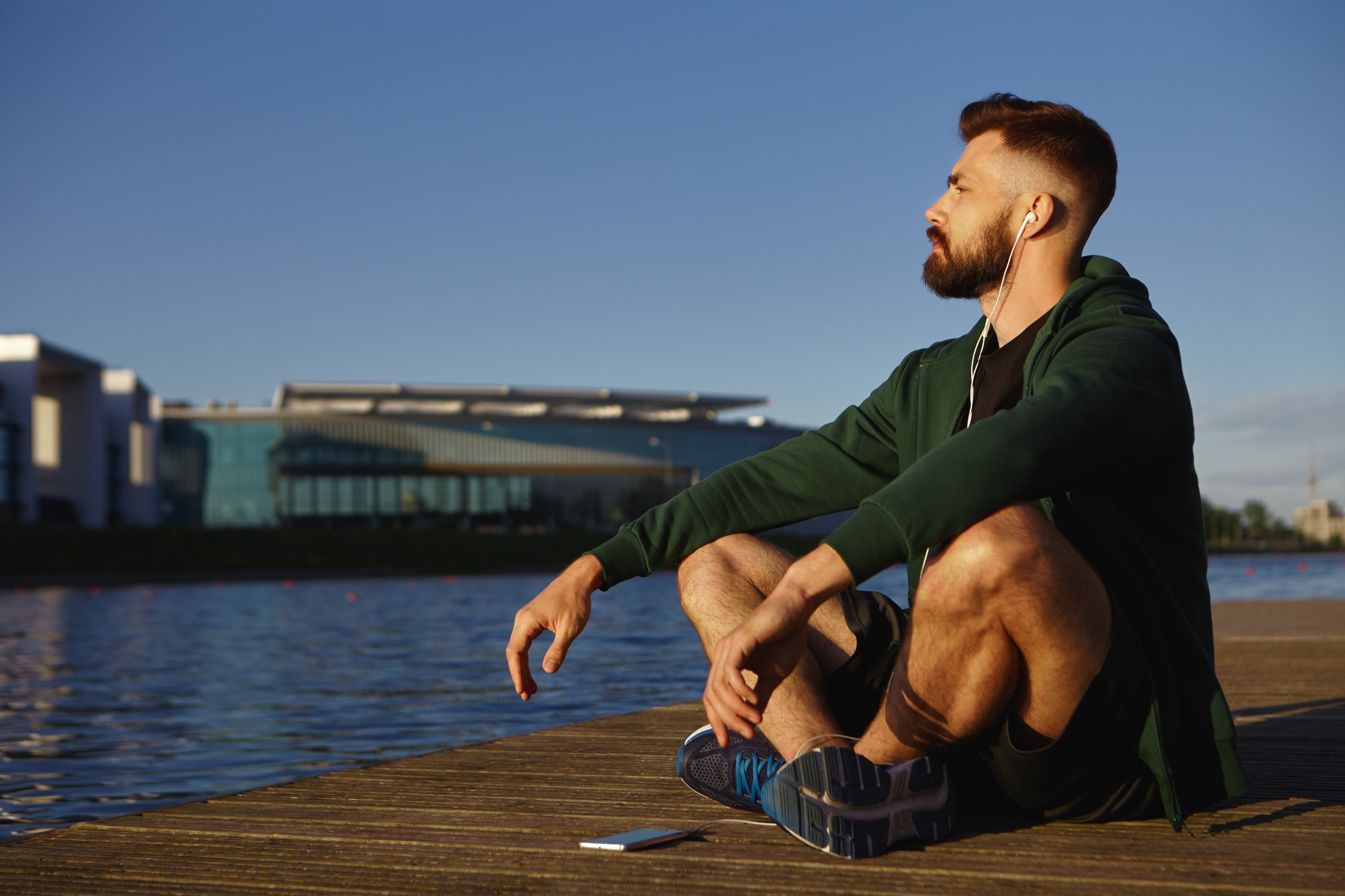 A young man sits in a meditative pose on a wooden dock by the water, practicing mindfulness with headphones at sunrise.