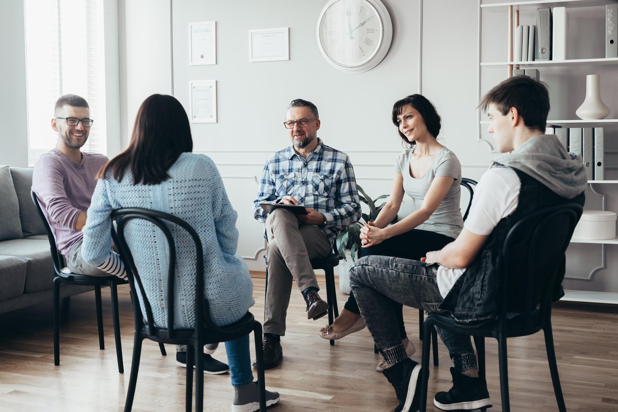 A diverse group of adults participates in a supportive group therapy program led by a male clinical professional.