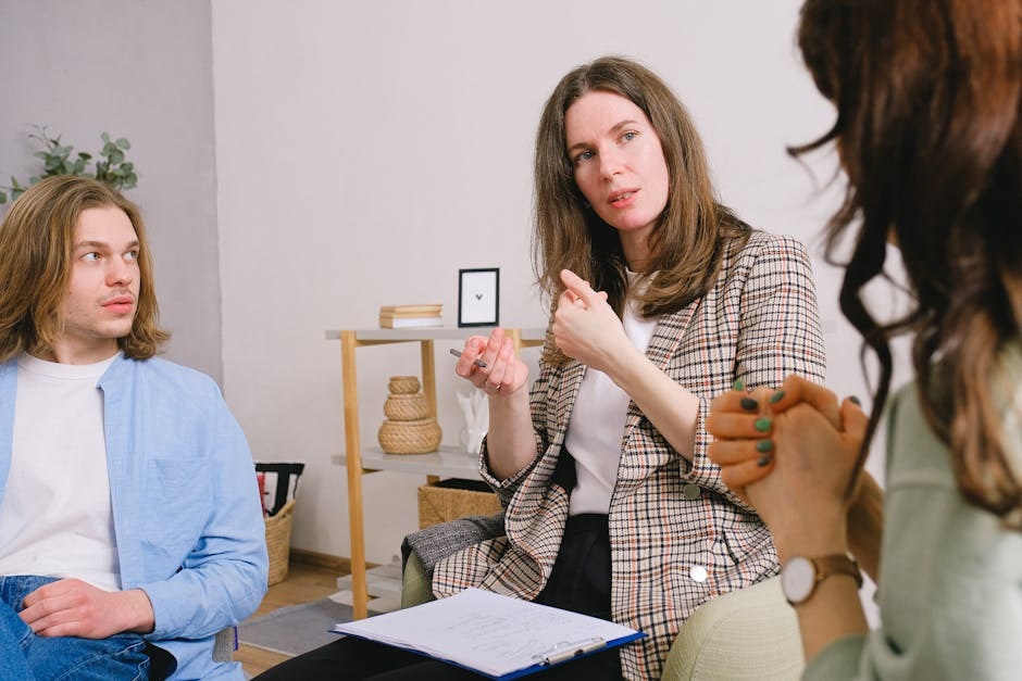 A female therapist in a checkered blazer leads a supportive DBT group session with two young adult participants.